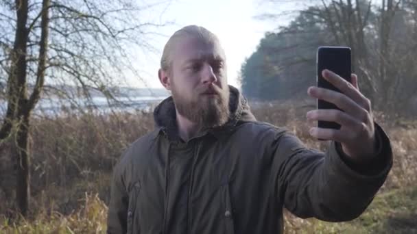 Sonbahar ormanlarında gölün önünde durup selfie çeken mutlu beyaz adamın portresi. Erkek turist fotoğraf çekmek için akıllı telefonunu kullanıyor. Yaşam tarzı, hobi, eğlence.. — Stok video