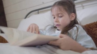 Yatakta kitap okuyan esmer, beyaz bir kızın yakın çekimi. Mutlu sevimli çocuk yatmadan önce hikayelerin tadını çıkarıyor. Hobi, yaşam tarzı, boş zaman.