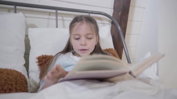 Πορτρέτο του όμορφου καυκάσιου κοριτσιού ξαπλωμένη στο κρεβάτι και διαβάζοντας βιβλίο. Έξυπνο παιδί που πάει για ύπνο το βράδυ. Χρόνος ύπνου, χόμπι, αναψυχή. — Αρχείο Βίντεο