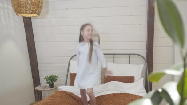 Middle shot of happy little Caucasian girl jumping on bed and smiling. Joyful brunette child having fun indoors on weekends. Leisure, joy, happiness. — ストック動画