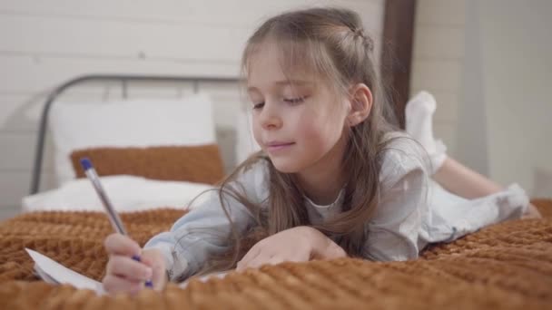 Portret van een blank meisje dat op bed ligt en schrijft. Intelligent kind dat laat zien dat ik een ideeëngebaar heb en haar gedachten noteert. Lifestyle, ontspanning, hobby. — Stockvideo