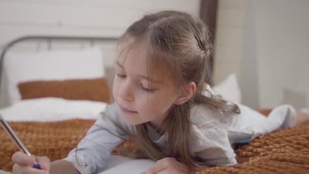 Kamera nähert sich dem niedlichen Gesicht eines kleinen kaukasischen Mädchens, das auf dem Bett liegt und schreibt. ziemlich fröhliches Kind, das in die Kamera schaut und den Finger schüttelt. Kindheit, Freude, Freizeit. — Stockvideo