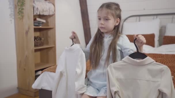 Portret małej białej dziewczynki siedzącej na łóżku z dwoma wieszakami i myślącej. Ładne dziecko wybiera strój rano. Styl życia, moda. — Wideo stockowe