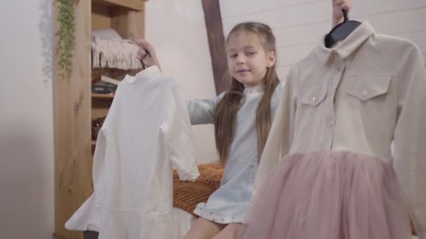 Schattig blank brunette meisje kiezen outfit voor school in de ochtend. Mooie dame die twee hangers vasthoudt, er een selecteert en het schot achterlaat. Lifestyle, mode, kindertijd. — Stockvideo