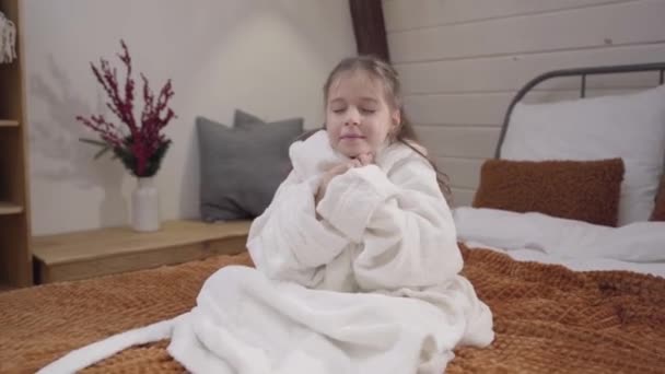 Camera approaching to charming Caucasian girl with beautiful eyes sitting on bed dressed in white bathrobe. Portrait of cute little lady after shower in bedroom. Happiness, lifestyle, leisure. — ストック動画