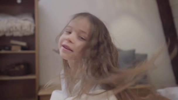 Retrato de cerca de una linda niña morena sacudiendo el pelo largo. Sonriente niño feliz sentado en la cama, mirando a la cámara y sonriendo. Ocio, felicidad, estilo de vida. Movimiento lento . — Vídeo de stock