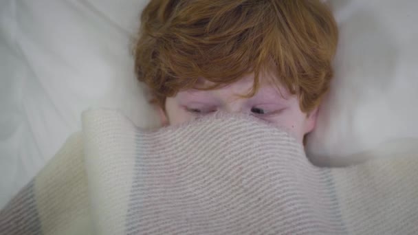 Primer plano de los ojos del pelirrojo caucásico con la cara cubierta de manta. Chico pequeño acostado en la cama y mirando a su alrededor. Infancia, ocio, diversión . — Vídeos de Stock