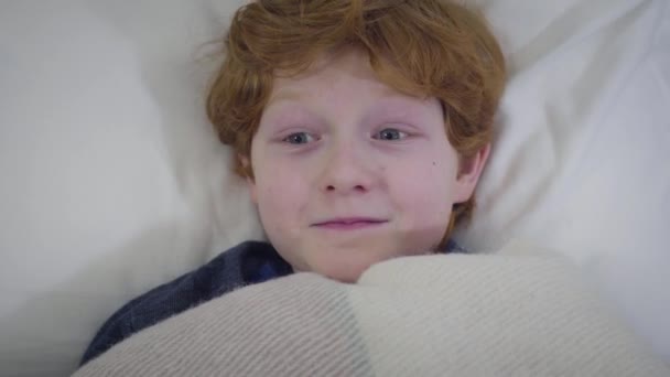 Portret van een vrolijk blank roodharig kind dat in bed ligt en zich verstopt onder een deken. Gelukkige jongen die 's avonds gaat slapen. Vreugde, vrije tijd, levensstijl. — Stockvideo