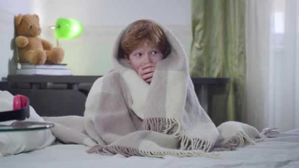 Retrato de lindo niño caucásico sentado en la cama cubierto con manta y mirando a su alrededor. Niño asustado con el pelo rojo dejado solo en casa. Infancia, soledad, miedo . — Vídeos de Stock