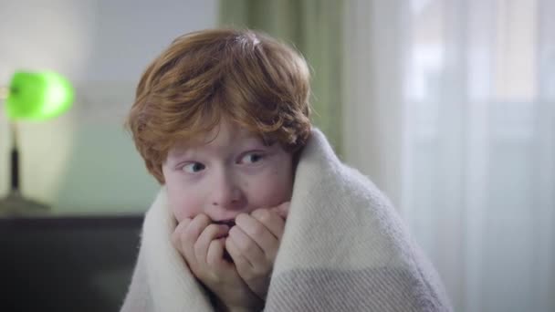 Portrait of terrified Caucasian redhead boy sitting indoors covered with blanket and looking around. Little child left home alone. Childhood, fear, lifestyle. — Stock Video