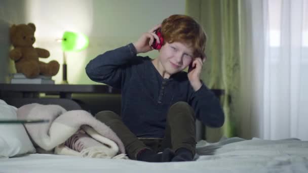 Enthousiaste blanke roodharige jongen in oortjes die naar muziek luistert. Vrolijke kleine man schudt hoofd als zittend op bed thuis. Hobby, levensstijl, kindertijd. — Stockvideo