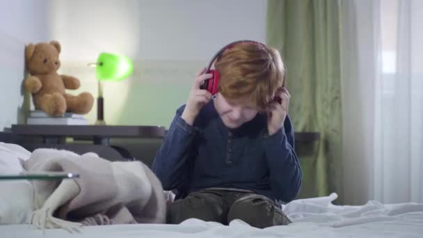Μικρό Καυκάσιο αγόρι με κόκκινα μαλλιά που φοράει ακουστικά και κουνάει το κεφάλι συναισθηματικά. Χαριτωμένο παιδί που ακούει ροκ μουσική σε εσωτερικούς χώρους. Χαρά, χόμπι, τρόπος ζωής. — Αρχείο Βίντεο