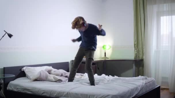 Actieve roodharige jongen die op bed springt en naar muziek luistert. Vrolijk blank kind heeft plezier thuis in het weekend. Vrije tijd, geluk, kindertijd. — Stockvideo