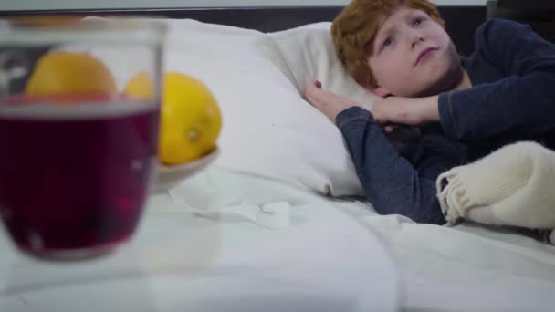 Ill Caucasian little boy lying in bed at the background and turning his back to camera. Hot drink and oranges placed on the table at the foreground. Healthcare, sickness, illness. — 비디오