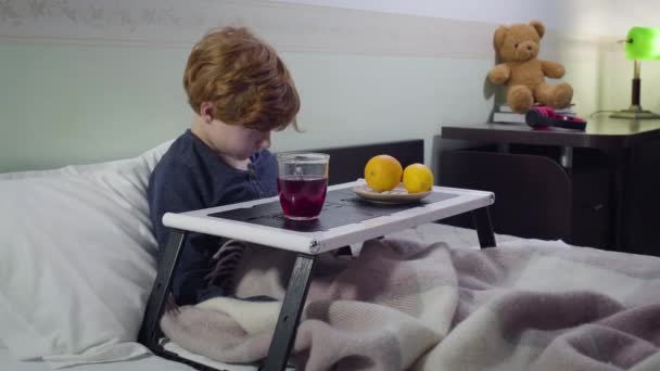 Triste pelirrojo caucásico acostado en la cama y bebiendo té de frutas calientes. Retrato del niño enfermo alojado en el dormitorio con bandeja de la cama. Salud, medicina, enfermedad . — Vídeo de stock