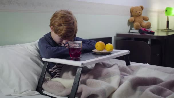 Menino caucasiano doente perturbado sentado na cama em frente à bandeja com bebida quente e laranjas. Retrato de criança ruiva bonito triste ficar em casa. Cuidados de saúde, medicina, doença . — Vídeo de Stock