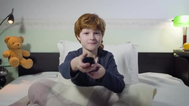 Vue de face portrait de mignon garçon rousse caucasien assis sur le lit et changer de canaux avec télécommande. Charmant enfant mâle à la recherche de dessins animés intéressants. Enfance, loisirs, coucher . — Video