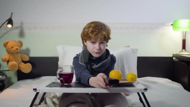 Portrait of ill Caucasian boy switching channels with remote control and watching TV. Cute redhead child sitting on bed in front of tray with tea and oranges. Sickness, healthcare, lifestyle. — 비디오