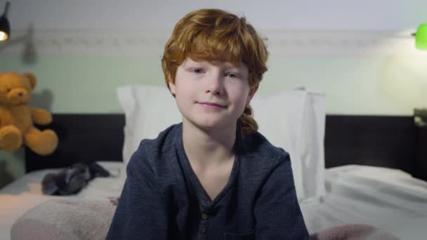 Närbild porträtt av söt vit pojke med rött hår och grå ögon tittar på kameran och ler. Charmigt litet barn poserar i sovrummet hemma. Barndom, lycka, fritid. — Stockvideo