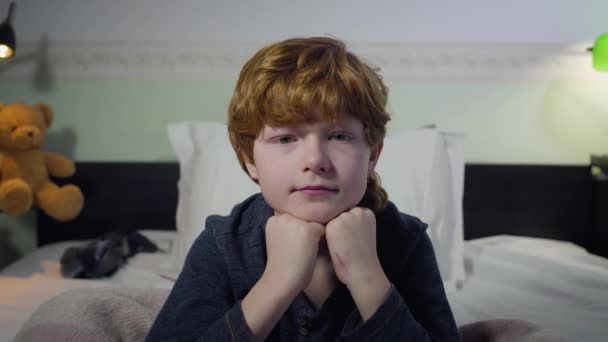 Huvudskott av söta kaukasiska rödhåriga unge sitter på sängen och tittar på kameran. Porträtt av en liten pojke i sovrummet hemma. Livsstil, barndom. — Stockvideo