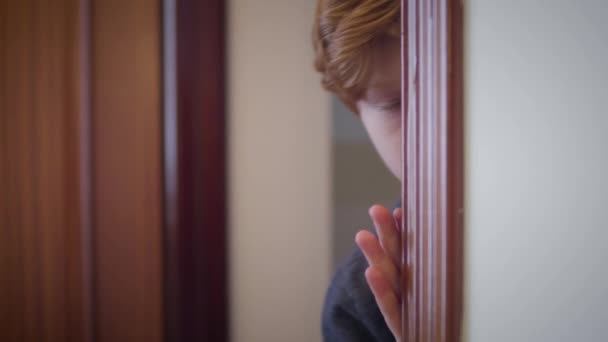 Close-up of cute redhead Caucasian boy looking out from behind doorway and hiding back. Portrait of scared little kid indoors. Fear, stress, childhood. — Stock Video
