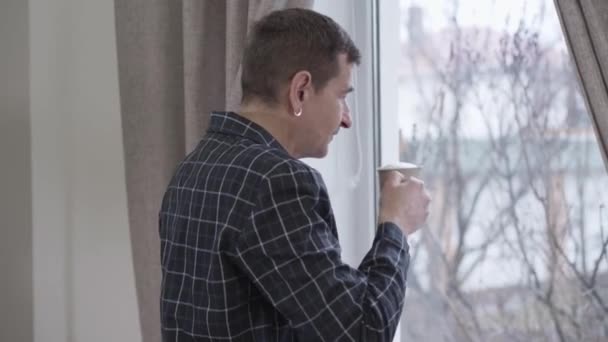 Back side view of adult Caucasian man with earring drinking coffee in the morning in front of window. Middle aged man in pajamas enjoying morning cappuccino and waving. Joy, happiness, lifestyle. — стокове відео