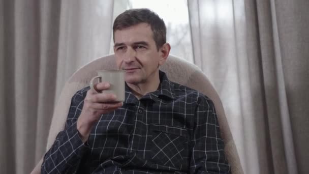 Retrato de hombre caucásico de mediana edad positivo sentado en sillón y bebiendo café. Hombre adulto guapo pasando la mañana en casa. Ocio, felicidad, estilo de vida . — Vídeos de Stock