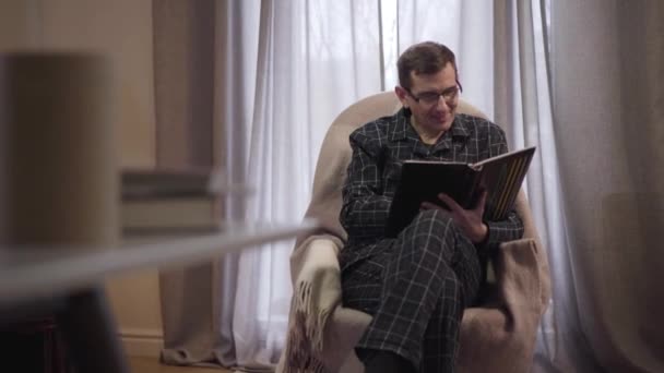 Portrait d'un homme caucasien adulte souriant en tenue de nuit assis dans un fauteuil à la maison et regardant des photos dans un album photo. Heureux homme d'âge moyen se reposant seul à la maison le soir . — Video