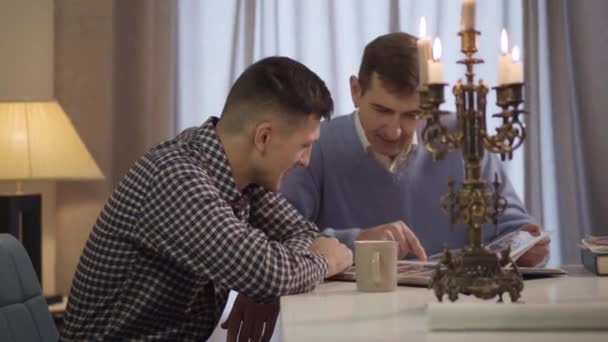 Side view of adult handsome Caucasian man sitting at the table and talking with middle aged father. Son and parent spending evening indoors chatting. Lifestyle, happiness, leisure. — Stock Video