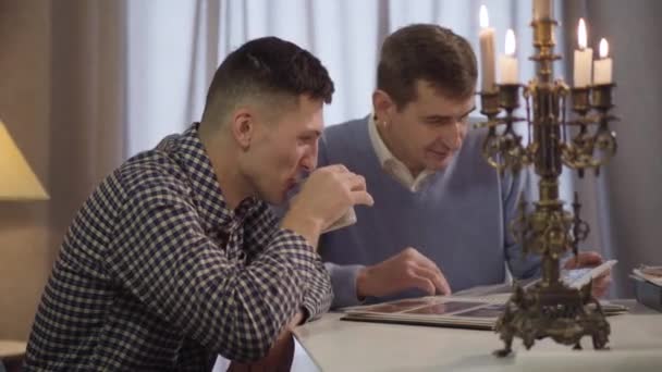 Amistoso padre caucásico e hijo bebiendo café y charlando por la noche en casa. Dos hombres adultos sonrientes hablando y mirando fotos en el álbum de fotos en el interior. Vinculación, familia, estilo de vida . — Vídeos de Stock