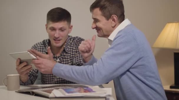 Vista lateral de hombre caucásico sonriente de mediana edad usando tableta y hablando con hijo adulto en casa. Hombres alegres descansando juntos en el interior por la noche. Ocio, estilo de vida, familia . — Vídeo de stock