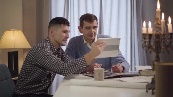 Retrato de vista lateral del hombre caucásico adulto sonriente haciendo videollamada para su padre de mediana edad. Feliz hijo y papá usando la tableta en el interior de la noche. Tecnologías modernas, estilo de vida, generaciones . — Vídeos de Stock