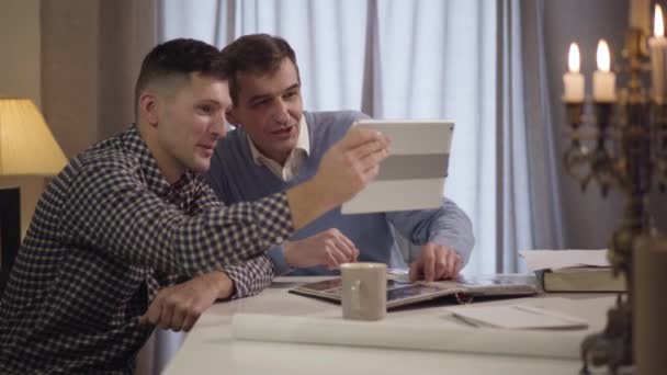 Dois homens adultos caucasianos usando tablet e acenando na tela. Pai e filho fazendo videochamada à noite em casa. Família, gerações, tecnologias modernas . — Vídeo de Stock