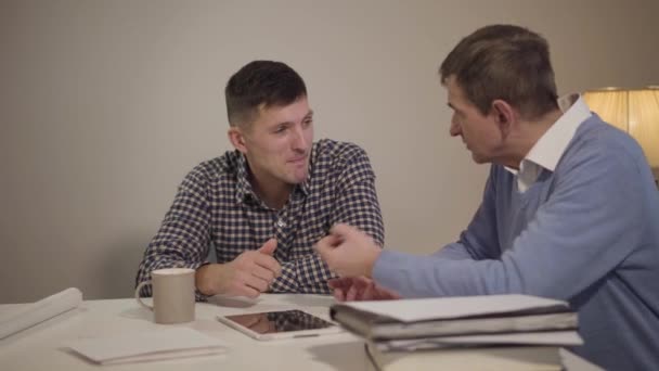 Twee blanke mannen zitten aan tafel en praten. Serieuze vader en zoon bespreken iets binnen. Verbinden, familie, levensstijl. — Stockvideo