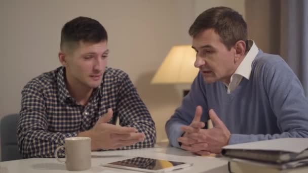 Portrét dospělého kavkazského syna a otce středního věku, jak si večer doma povídají. Dva muži sedí u stolu a povídají si. Rodina, generace, životní styl. — Stock video