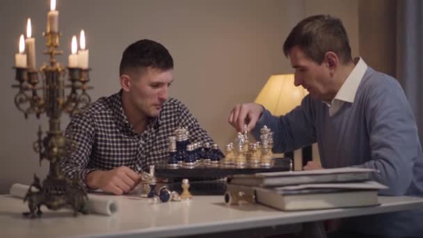 Cheerful Caucasian son and father playing chess in the evening at home. Adult man spending free time with parent indoors. Bonding, happiness, lifestyle, hobby. — 비디오