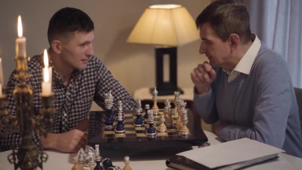 Portrait of intelligent middle aged Caucasian man teaching his adult son to play chess. Two men spending evening at home. Lifestyle, hobby, intelligence. — 비디오