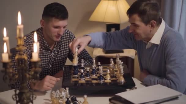 Two positive Caucasian men competing in chess play. Father and son sitting at the table in the evening. Hobby, lifestyle, family. — Stock Video