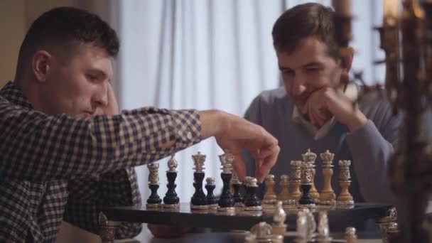 Gros plan d'hommes caucasiens confiants jouant aux échecs à l'intérieur. Changement d'orientation du fils adulte au père d'âge moyen. Des échecs concentrés passent la soirée à la maison. Loisirs, passe temps, mode de vie . — Video