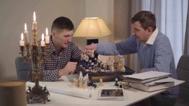 Cute Caucasian brunette boy with curly hair coming to grandfather and dad playing chess. Multi-generation family spending evening at home together. Hobby, lifestyle, leisure. — 비디오