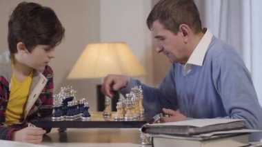 Büyükbabamla kapalı mekanda satranç oynayan beyaz çocuğun yan görüntüsü. Torun hafta sonlarını büyük anne ve büyükbabasıyla geçiriyor. Hobi, yaşam tarzı, boş zaman.