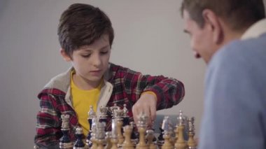 Satranç oynayan ve zafer jesti gösteren konsantre olmuş beyaz çocuk portresi. Ön planda bulanık dedesi elini sıkarken. Zeki çocuk yarışmayı kazanıyor. Yaşam tarzı, hobi.