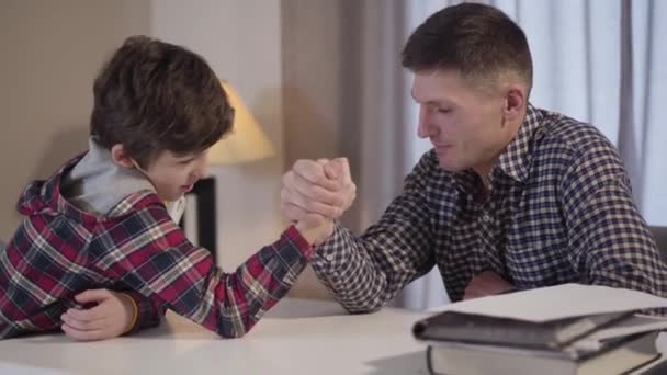 Close-up side view of Caucasian father and son competing in arm-wrestling. Sad kid losing, parent cheering him up. Lifestyle, leisure, family. — Stock Video