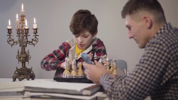 Portrait of cute smart Caucasian boy placing chess on board and talking to father sitting at the foreground and using smartphone. Son asking attention from parent in the evening. Family, relationship. — Stock Video