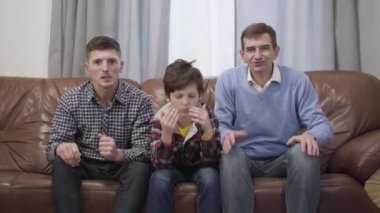Portrait of three men of different ages watching football match on TV and gesturing. Sad Caucasian son, father and grandfather discussing their team losing. Leisure, family, lifestyle.