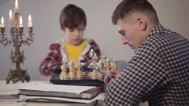 Akıllı telefonu arka planda satranç tahtasının önünde oturan üzgün oğlu olarak kullanan beyaz bir adamın yan görüntüsü. İletişim sorunları, ebeveynlik, yaşam tarzı. — Stok video