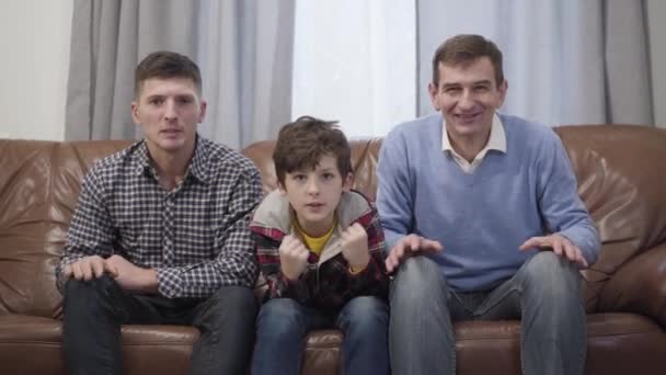 Портрет кавказской семьи футбольных болельщиков, смотрящих матч по телевизору. Мужчина средних лет, его взрослый сын и маленький внук делают победный жест, когда их команда выигрывает. Хобби, досуг . — стоковое видео