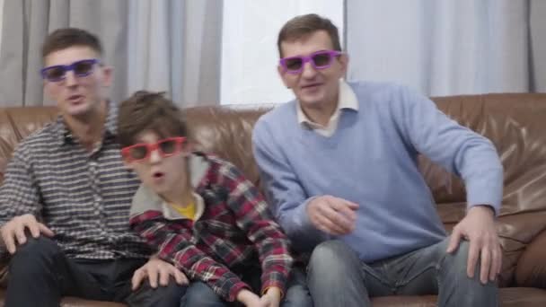 Three Caucasian men of different ages sitting on couch at home and watching TV in 3D goggles. Cheerful multi-generation family having fun indoors on weekends. Joy, leisure, lifestyle. — Stockvideo