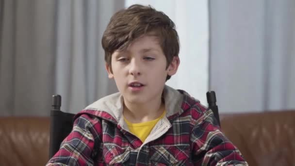 Retrato de lindo niño caucásico con el pelo rizado morena y ojos grises sentado en el interior y hablando a la cámara. Niño con ropa casual posando en casa. Estilo de vida, ocio, alegría . — Vídeo de stock