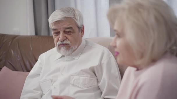 Egy közeli kép az öreg fehér hajú férfiról, aki otthon beszélget a feleségével. Házas nyugdíjasok, akik együtt pihennek esténként a házban. Kötődés, támogatás, szeretet. — Stock videók